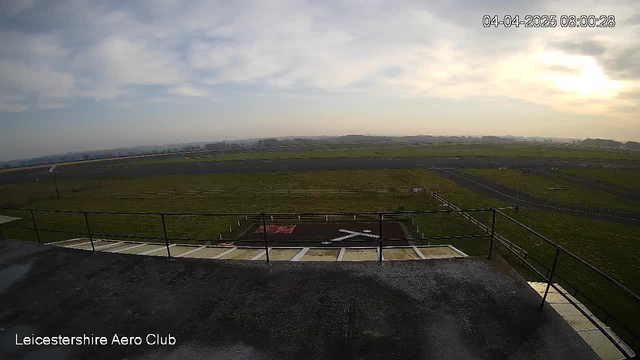 A wide view of an open field belonging to the Leicestershire Aero Club, featuring a runway stretching into the distance. The foreground shows a low railing and a tarmac area with a large white 'X' marking, indicating a landing or take-off zone. The sky is partly cloudy with a soft, diffused light, suggesting early morning. Green grass and a few scattered barriers are visible, while the horizon features gentle rolling hills.