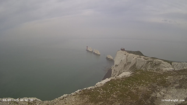 A panoramic view of a calm sea under a light gray sky. In the foreground, there are grassy cliffs with white rock formations. Several large, white rocky outcrops are visible offshore, with one featuring a small red lighthouse. The water is blue-gray, and there's a faint horizon line in the distance. The scene appears serene and slightly overcast.