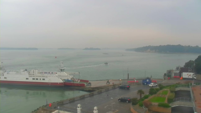 A cloudy harbor scene featuring a large white and red ferry boat docked at the pier. In the foreground, there are several parked cars and a small blue building. A smaller boat can be seen moving away from the ferry towards the calm water. The background includes a hazy landscape of land and trees lining the shoreline, with a few distant islands visible in the water. The atmosphere is overcast, creating a muted color palette.