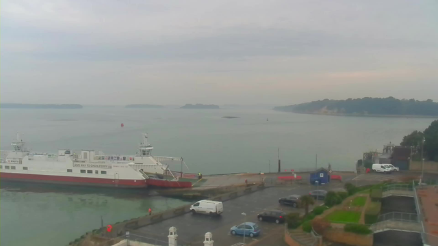 A ferry with a white and red hull is docked at a quay, with a calm bay visible in the background. The water is reflecting the overcast sky, which has a grayish hue. Several islands are visible in the distance, and a red buoy is floating in the water. In the foreground, there is a parking lot with several cars, including a white van, and some green landscaping nearby. The scene is tranquil, with no signs of movement on the water.