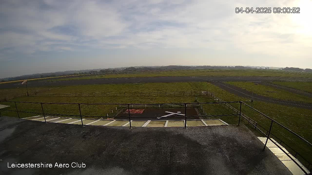 A view from a high vantage point overlooking an airfield. In the foreground, there is a flat, gray rooftop surface with some white markings. Below, a grassy area is visible, bordered by a wooden fence. The airfield is characterized by a long, dark runway and several small white markers. In the distance, a low horizon with green fields and a partly cloudy sky can be seen. The time and date displayed on the image indicate it is early morning on April 4, 2025.