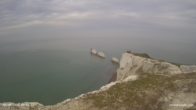 A coastal scene featuring white chalk cliffs rising sharply from a calm, greenish-blue sea. In the distance, two rock formations emerge from the water. One has a red and white lighthouse structure on it, while the other is a group of larger, white rocks. The sky is mostly cloudy, with a light grayish tone. The foreground has grassy patches and some rocky terrain. The date and time are displayed in the corner.