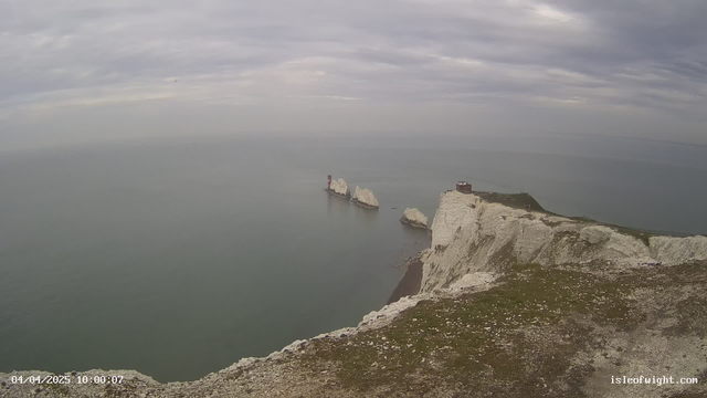 A coastal landscape featuring white chalk cliffs that drop down to a calm, grayish-blue sea. In the distance, several rocky formations rise from the water, including two narrow outcrops topped with buildings, one of which is painted red. The sky is overcast with soft gray clouds, and the foreground displays grassy areas interspersed with pebbles. The scene conveys a serene, natural environment on a cloudy day.