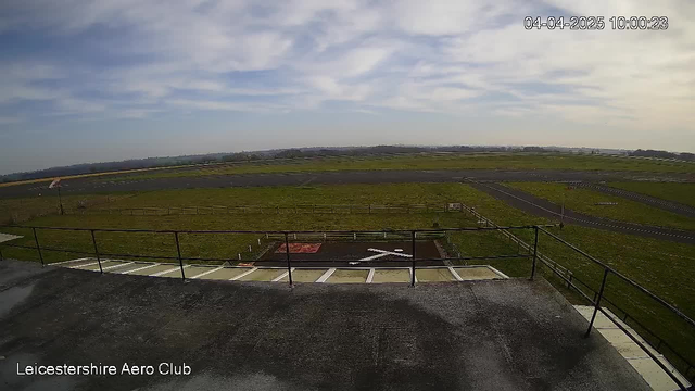 A wide view of an airfield with a grassy runway and a helicopter landing pad featuring a white cross in the center. The scene includes a clear blue sky with some scattered clouds. In the distance, there are rolling hills, and the foreground shows a balcony railing. The area is partially enclosed by a fence, indicating the boundaries of the airfield.