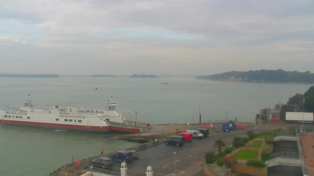 A view of a calm body of water with several small islands in the distance. In the foreground, a white ferry with a red lower hull is docked at a terminal. Nearby, there are a few cars parked in a lot, including a red van and a dark vehicle. The scene is under a cloudy sky with soft, muted colors. The shoreline features greenery and a pathway leading to the water's edge.