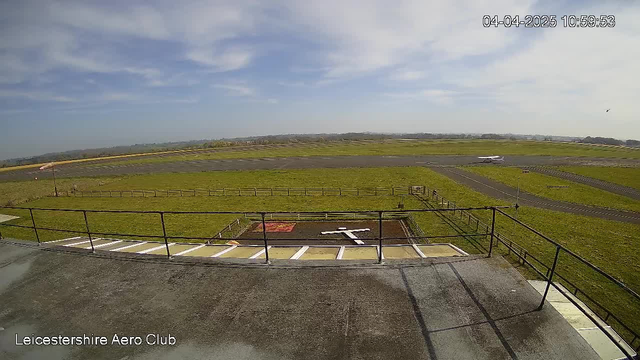A view from a high vantage point overlooking an airfield. The foreground shows a rooftop railing and surface, with a large white cross marked on the ground below. In the middle background, a green field stretches out, framed by a perimeter fence. A runway is visible, leading towards a distant horizon where small aircraft can be seen. The sky is mostly clear with some clouds, indicating a bright day.
