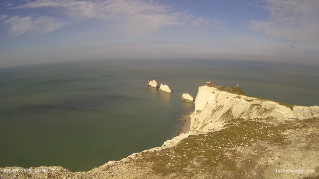 A coastal landscape is depicted with white chalk cliffs on the right side of the image. Two small lighthouse structures, one red and white and the other white, are visible on the rocks extending into the calm sea. The water is a gradient of green and blue, reflecting the clear sky above, which is partly cloudy. The left side of the image shows a grassy area near the cliff's edge. The scene conveys a tranquil coastal environment.