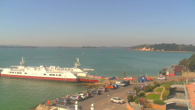 A calm waterfront scene featuring a white ferry docked at a harbor. The ferry is partially out of view, with the text "Give Way to Chain Ferry" visible on its side. The water is a mix of blue and green hues, with a few boats in the distance. The shoreline is visible in the background, lined with trees and sandy cliffs. In the foreground, a parking lot filled with cars in various colors, with some people walking nearby. The sky is clear and bright.
