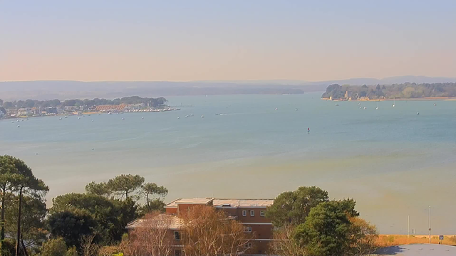 A scenic view of a calm bay with a light blue-green water surface. Multiple small boats are visible on the water, some anchored and others in motion. In the background, there is a shoreline with trees and buildings, including a prominent structure that looks like a castle or historic site. The horizon features rolling hills, and the sky is clear with a slight haze, suggesting a warm, sunny day. The foreground includes areas of vegetation and a multi-story building partially obscured by trees.