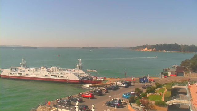 A ferry is docked at a waterfront, with vehicles parked nearby. The water is a vibrant blue-green, and small waves are gently lapping against the shore. In the distance, there are hills with trees and a sandy beach, while a few boats can be seen on the water. The sky is clear and bright. The area features landscaped greenery and pathways leading down to the water's edge, with people walking near the ferry and along the promenade.