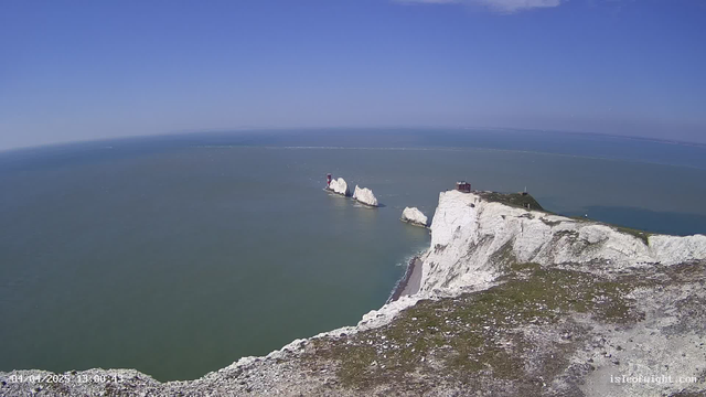A panoramic view of a coastline featuring steep white cliffs that drop into the sea. In the background, the ocean is a calm blue, with a clear sky above. Two small lighthouses are visible on the cliffs, and there’s a hint of a path leading along the shore. The scene is bright and sunny, showcasing a natural landscape with a mix of water and rocky terrain.