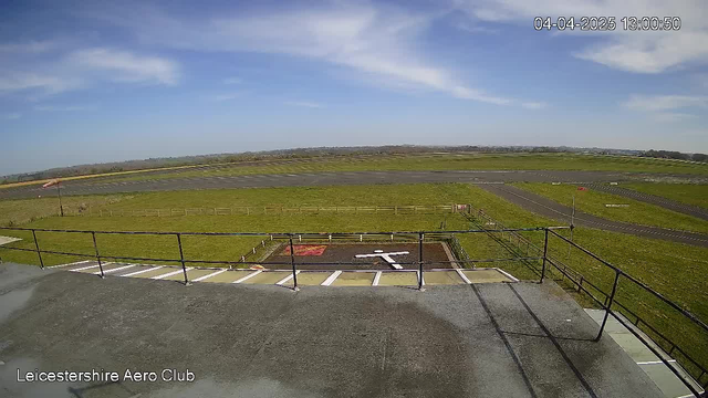 A clear blue sky with a few wispy clouds stretches above a grassy airfield. In the foreground, a flat surface with white markings is visible alongside a helipad symbol. The airfield is bordered by a wooden fence, and the runway extends into the background, where more grass and a distant landscape can be seen. The location is identified as Leicestershire Aero Club, and the time is noted in the corner of the image.