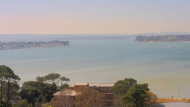 A panoramic view of a calm bay with boats scattered on the water. In the foreground, there are trees and a low building, while further in the distance, there are more boats and a shoreline lined with houses. The sky is clear, and the background features distant hills. The overall scene conveys a serene and tranquil atmosphere.