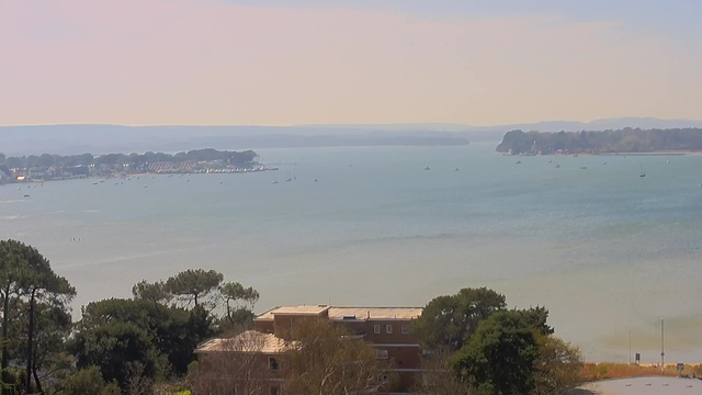 A scenic view of a calm body of water with small boats scattered across it. In the foreground, there are trees and a partially visible building. The shoreline features houses and a distant peninsula with greenery. The sky is bright with a light haze, indicating daytime, and gentle hills are visible in the background.
