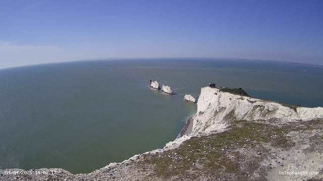 A panoramic view of a coastline on a clear day. In the foreground, there are steep, chalky white cliffs with some green vegetation. The ocean stretches out in the background, reflecting the blue sky with a few clouds. A series of rocky outcrops jut out into the water, with two small red structures visible on them. The horizon line is mostly straight, indicating calm seas. The image captures a sense of tranquility and natural beauty.