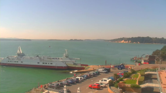 A view of a harbor with a large ferry docked at the shore, alongside a variety of parked cars in the foreground. The water is calm and blue, with gentle waves. In the background, there are green hills and a sandy coastline. A few boats can be seen in the distance. The sky is clear with a few clouds. There are also plants and walkways near the water's edge.