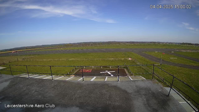 A clear sky with a few wispy clouds is visible above a green landscape. In the foreground, there is a dark surface with a white painted cross and a red square at a small aerodrome. A level area of grass and a paved runway stretch across the background. The runway is marked with yellow lines and the horizon shows distant hills and trees. A metal railing runs along the front edge of the viewing area.