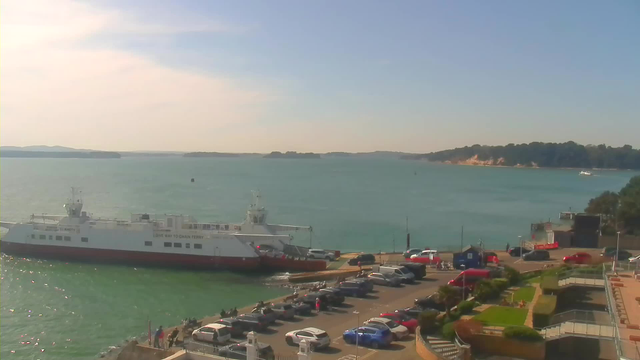 A view of a harbor with a large white ferry docked on the left side. The ferry has a red stripe at the bottom and is labeled with "GIVE WAY TO CHAIN FERRY." In the foreground, there is a parking lot filled with various cars, and a few people are gathered near the water's edge. The ocean is calm and blue, and in the background, there are green hills and a sandy coastline under a bright sky. The overall scene conveys a sunny, peaceful day by the water.