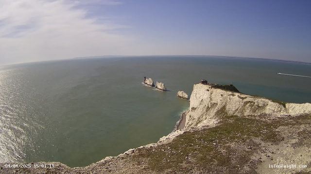 A panoramic view of a coastal landscape featuring white chalk cliffs along the shoreline. In the distance, several rocky outcrops rise from the sea, with a small building visible on one of them. The ocean appears calm, reflecting the blue sky, which is lightly clouded. Below the cliffs, there is a narrow beach, and a boat can be seen moving through the water closer to the horizon. The scene conveys a serene coastal atmosphere.