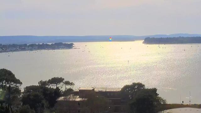 A serene view of a body of water under soft lighting. The surface reflects shimmering light, creating a silvery appearance. Several small boats are visible, scattered across the water. In the distance, a shoreline with trees and buildings can be seen, along with hills fading into the background. The overall atmosphere is calm, with a light breeze suggested by the gentle waves.