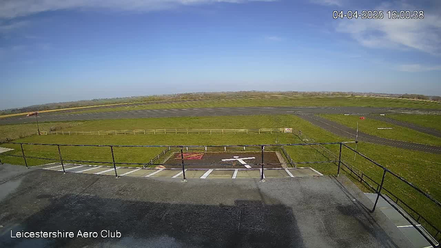 A view from a webcam shows a wide, open field at the Leicestershire Aero Club. In the foreground, there is a flat rooftop area with a railing. Below, a grass airstrip extends into the distance, bordered by a wooden fence. A white cross marking for helicopter landings is visible on the ground, along with areas marked in red and white. The background features a clear blue sky with a few scattered clouds, and gentle hills can be seen far off in the distance. The time is displayed on the top right corner of the image.