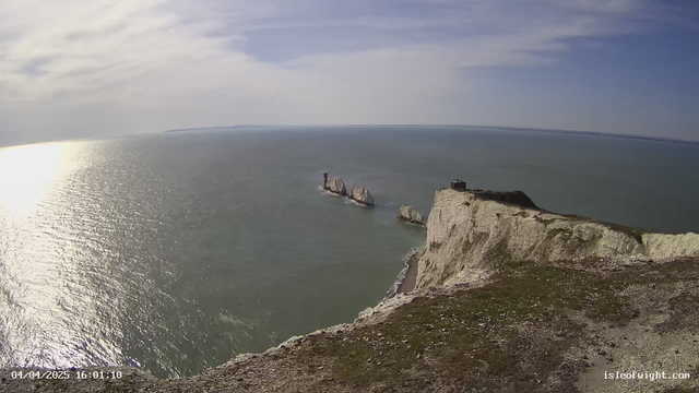 A panoramic view of the coastline, featuring white chalk cliffs under a partly cloudy sky. Below, the calm sea reflects sunlight, creating a shimmering effect. In the distance, a group of rocky formations emerges from the water, while a small structure sits atop one of the cliffs. The landscape is grassy, with sparse vegetation and pebbles visible on the surface. The date and time are noted in the bottom left corner.