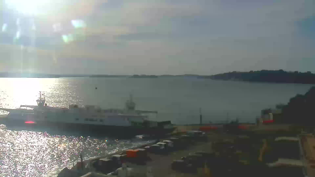 A wide view of a body of water under a partly cloudy sky. Sunlight reflects off the gently rippling surface of the water, creating a shimmering effect. On the left, a white ferry boat is docked near a harbor, with some vehicles parked nearby. In the background, there are distant hills and small islands on the horizon. The scene conveys a sense of calm and tranquility.