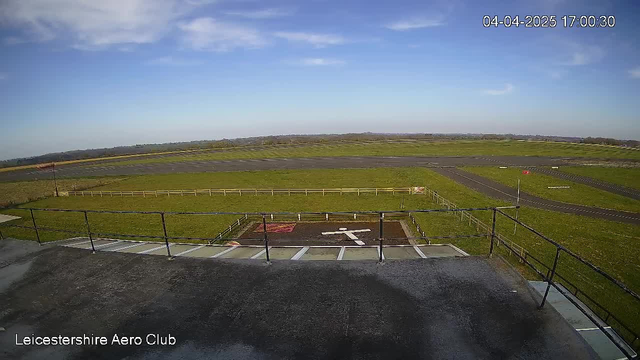 A wide view of an airport from a webcam, taken from a high vantage point. The image shows a grassy area with a runway in the distance, under a clear blue sky with some clouds. In the foreground, there is a boundary railing and markings on the ground, including a white figure symbolizing a person and a red square area. The runway stretches across the image, with a few lines indicating taxi paths and a windsock visible on the side. The date and time are displayed in the upper right corner.