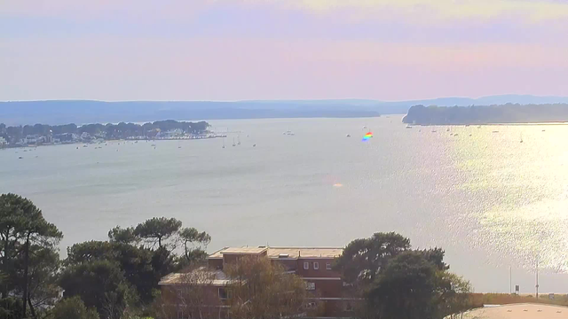 A serene view of a wide water body under a light sky, with gentle waves reflecting the sunlight. On the left, there are clusters of trees and buildings, while small boats can be seen dotting the water. In the distance, a shoreline lined with houses and a gentle slope of land is visible, with faint hills beyond. The scene conveys a peaceful, tranquil atmosphere.