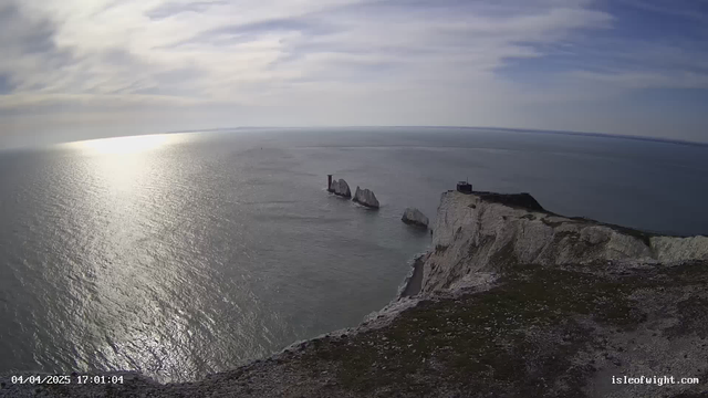 A panoramic view of a coastal landscape featuring high, white chalk cliffs on the right side of the image. The cliffs meet the sea, which reflects the soft light of the sun, creating a shimmering effect on the water's surface. In the distance, there are three prominent chalk rock formations rising from the water, with one of them showing the remains of a structure. The sky is partly cloudy, with a gentle gradient of blues and whites. The image captures a tranquil seaside scene on what appears to be a clear day.