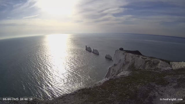 A scenic view of the ocean extending to the horizon under a mostly cloudy sky. The sun is low in the sky, casting a bright reflection on the water's surface, creating a shimmering effect. In the foreground, there is a grass-covered cliff edge with some rocky outcrops visible. To the right, white chalk cliffs are seen, and a small structure is situated at the top of the cliff. Three prominent rock formations rise from the water, with the nearest one displaying a vertical pillar. The overall atmosphere conveys tranquility and natural beauty.