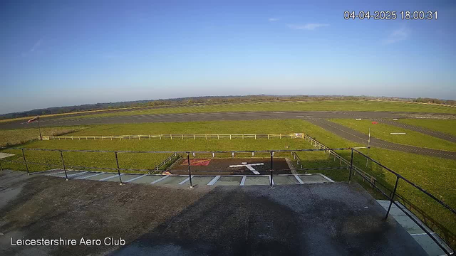 A clear blue sky is visible over a vast green landscape. In the foreground, there is a flat roof with a railing. Below, a fenced area includes a helipad marked with a white cross. To the right, asphalt runways are visible, with some vehicles or markers on them. The scene is well-lit, indicating daylight. The image is labeled with "Leicestershire Aero Club" in the lower left corner and shows the timestamp "04-04-2025 18:00:31" in the upper right corner.