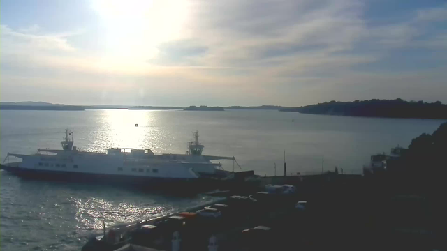 A bright, sunlit scene featuring a large white ferry docked at a harbor, with water shimmering under the sunlight. In the background, there are distant hills and a calm sea. Several parked cars line the harbor area, with more visible on the road leading to the waterfront. The sky is partly cloudy, and the sun is low on the horizon, casting a warm glow over the landscape.