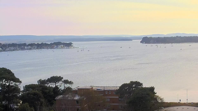 A scenic view of a calm body of water, with several small boats scattered across the surface. In the background, there are gentle hills and trees lining the shore. The sky features soft pastel colors, suggesting early morning or late evening light. In the foreground, there are some trees and a few buildings, partially obscured by greenery. The overall atmosphere is tranquil and serene.