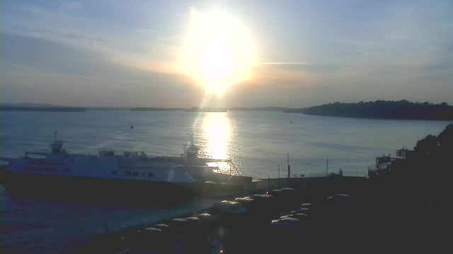A calm body of water reflects the bright sunlight as it sets on the horizon. A ferry boat is docked near the shore, partially obscured. Silhouettes of trees and hills are visible in the distance. A line of parked cars can be seen along the waterfront. The overall scene conveys a serene evening atmosphere with soft colors in the sky transitioning from blue to warm hues near the sun.