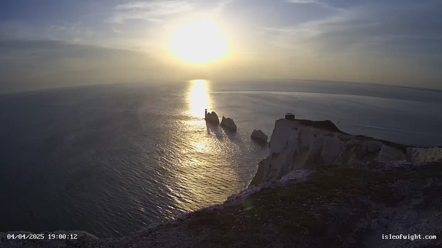 A scenic view of the ocean at sunset, with the sun low in the sky casting a golden reflection on the water. In the foreground, white chalk cliffs rise from the shore, leading to rock formations that jut out into the sea. The sky is a gradient of blue and soft orange with some clouds scattered. An observation tower is visible atop one of the cliffs, with the landscape extending into the distance.