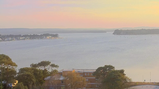 A serene view of a body of water during sunset, with the sky displaying soft pastel colors of pink, orange, and blue. In the foreground, there are trees and buildings. In the distance, small boats are scattered across the water, and a small marina can be seen along the shore, with hills gently rising in the background. The overall atmosphere is calm and tranquil.