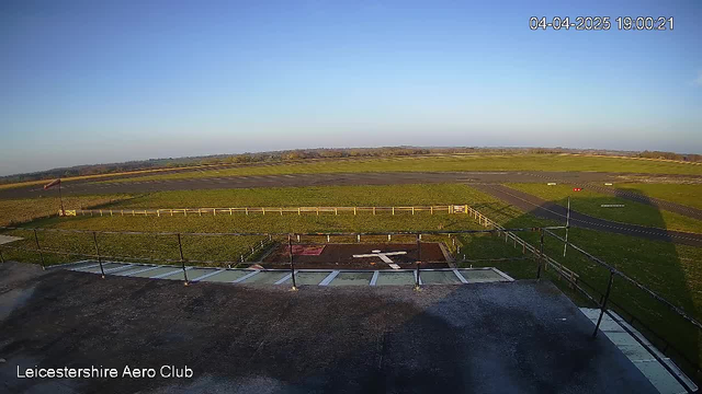 A clear sky during sunset with a gradient of blue to yellow hues. In the foreground, a helipad marked by a white cross sits on grass, surrounded by a fenced area. To the right, a paved runway extends into the distance, with grassy borders and a few scattered markers. The scene conveys a tranquil atmosphere at the Leicestershire Aero Club.