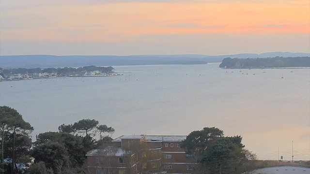 A panoramic view of a calm body of water at sunset. The sky displays soft hues of orange and pink, gradually fading into blue. In the foreground, there are trees and a few buildings. The water area is dotted with small boats, and in the distance, a shoreline with structures and trees is visible, with gentle hills beyond. The scene conveys a tranquil atmosphere.