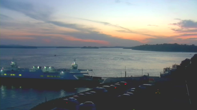 A view of a calm water body at sunset, with a blue ferry docked at a pier on the left. The sky is painted in soft hues of orange, pink, and blue, while shadows of distant islands are visible on the horizon. A row of parked cars is seen in the foreground, with some having their headlights on. The scene conveys a tranquil evening atmosphere.