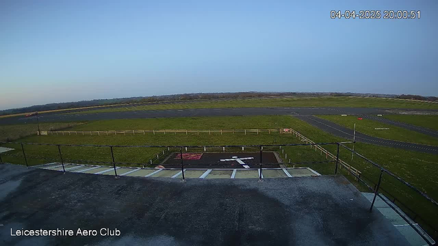 A view from a webcam at Leicestershire Aero Club, showing a clear blue sky above an open grassy area that serves as an airfield. In the foreground, there is a landing pad marked with a white cross. The area is surrounded by a wooden fence, and there are pathways visible on the ground leading towards the horizon. The time and date display shows April 4, 2025, at 20:00:51.