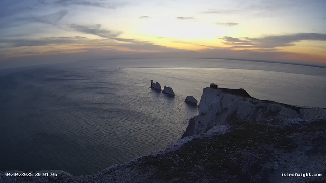 A view overlooking the ocean at sunset, with a serene skyline filled with soft clouds in shades of orange, pink, and blue. In the foreground, there is a rugged white cliff with some vegetation. Three distinctive rock formations rise from the water below, with a small lighthouse structure visible on the farthest rock. The water reflects the colors of the sky, creating a tranquil and scenic atmosphere. The bottom left corner displays a timestamp: "04/04/2025 20:01:06."