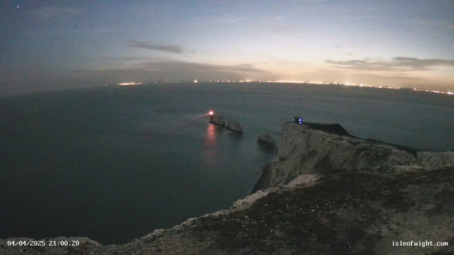A night scene from a webcam overlooking a dark sea. In the foreground, there is a rocky cliff with some vegetation. To the left, a lighthouse can be seen, emitting a red light that reflects on the water. The horizon shows faint lights, likely from a distant town or city. The sky is mostly clear with some scattered clouds and stars visible. The date and time at the bottom indicate it is April 4, 2025, at 9:00 PM.