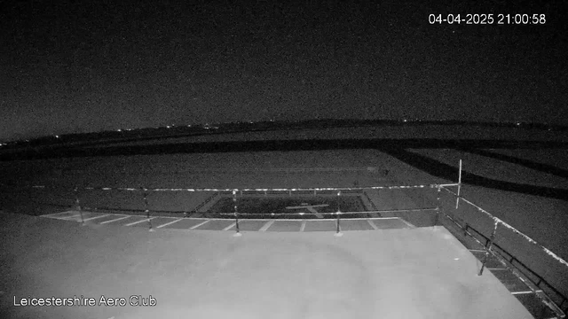 A nighttime view from a webcam showing a dark outdoor area, likely near an airfield. In the foreground, there is a railing with a horizontal bar, and a section of ground with what appears to be a helicopter landing pad, identifiable by a painted cross. The background features a flat landscape with distant lights or structures that create a faint glow against the dark sky. The time and date are displayed in the top right corner, indicating it is April 4th, 2025, at 9:00 PM.