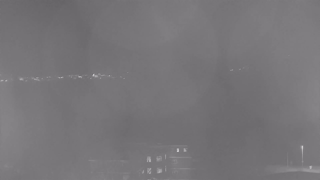 A dimly lit scene showing distant lights along a shoreline with a view of an urban area in the foreground. The image has low visibility and is primarily in shades of gray, with some areas appearing slightly clearer than others. There are indistinct outlines of buildings, with a few illuminated windows visible.