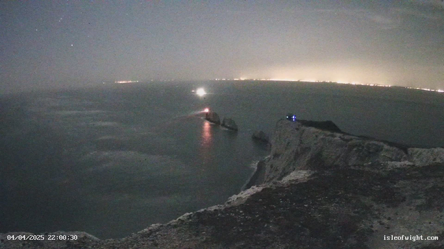 A nighttime coastal scene showing a cliff edge with rugged rocks jutting into the sea. In the distance, faint lights from a shoreline can be seen along with a beam of light from a lighthouse. The sky is dark with a few stars visible. The water appears calm, reflecting some of the ambient light.
