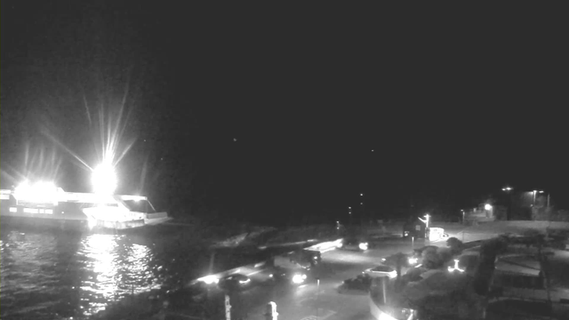 The image shows a nighttime scene featuring a dock area. A brightly lit ferry is docked on the left side, reflecting light on the water's surface. Several vehicles, including trucks and cars, are parked and moving around a circular area on the right. Streetlights illuminate the surroundings, providing some visibility in the dark environment. The background is mostly dark with limited visible details.
