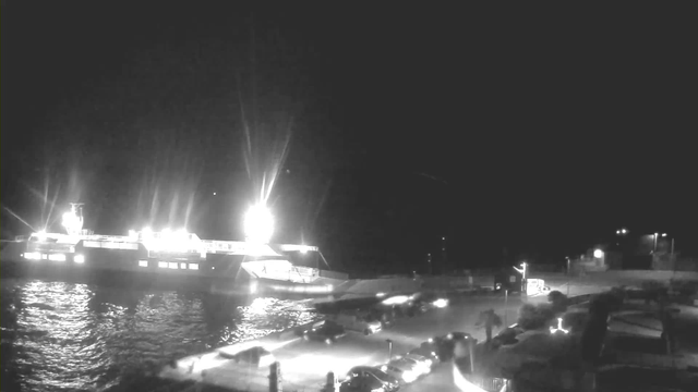 A nighttime waterfront scene featuring a large ferry boat docked at a pier, illuminated by bright lights. The water reflects the lights from the ferry, creating a shimmering effect. In the foreground, several parked cars are visible, along with a few distant lit structures along the shore. The sky is dark, with faint star visibility.