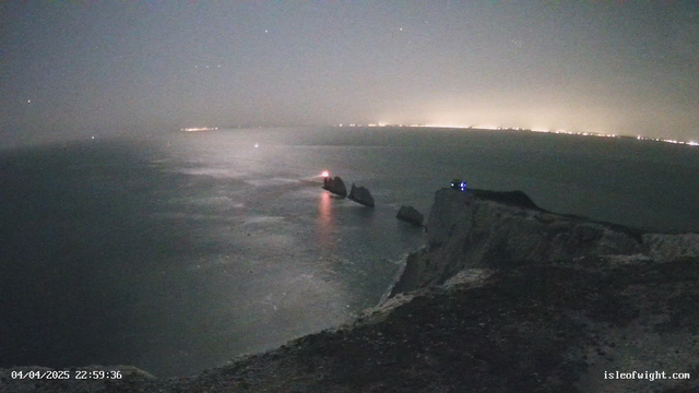 A nighttime view of a rocky coastline with gentle waves lapping against the shore. A lighthouse with a red light is visible on the left, while several jagged rocks are dotted along the water. In the distance, faint lights from a coastal town are noticeable against the dark sky. The scene is shrouded in a light mist, giving it a hazy appearance. The foreground shows a cliffside with a rugged texture. The timestamp in the corner indicates it is April 4, 2025, at 10:59 PM.