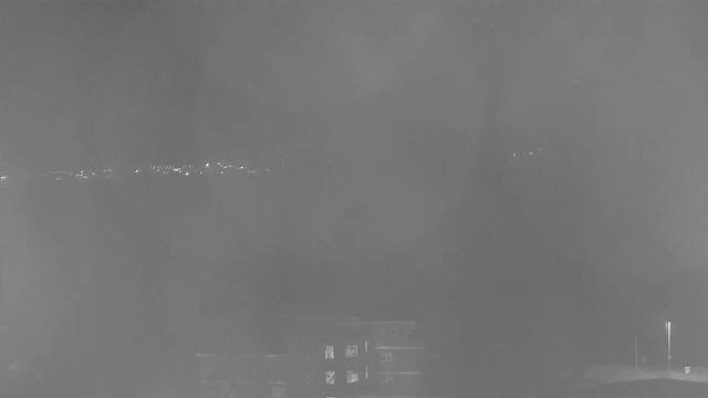 A foggy or hazy scene captured from a webcam, showing blurred outlines of lights in the distance, likely from buildings or streetlights. The foreground contains indistinct shapes, suggesting structures, but overall visibility is very low due to the haze. The image is primarily in shades of gray.